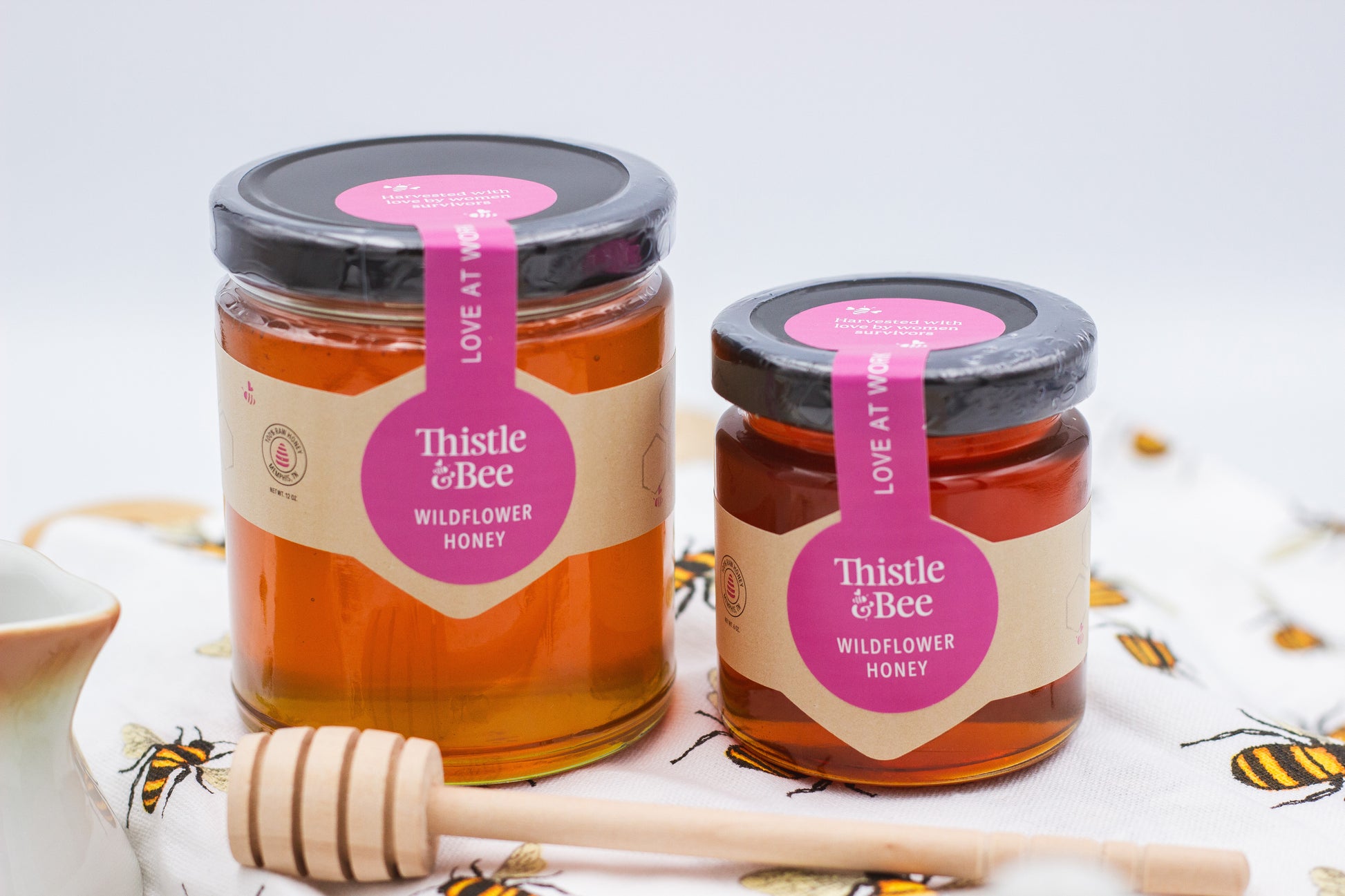 Two jars of Thistle & Bee wildflower honey with a honey dipper on a white background.