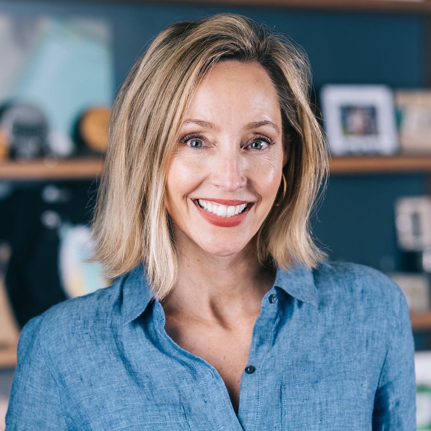 A smiling blonde woman wearing a blue button-up shirt, photographed in a warm indoor setting with blurred shelves in the background.